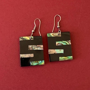 May include: A pair of rectangular black earrings with silver hooks. The earrings feature inlaid iridescent abalone shell accents in shades of green, blue, and silver. The earrings are displayed against a red background.