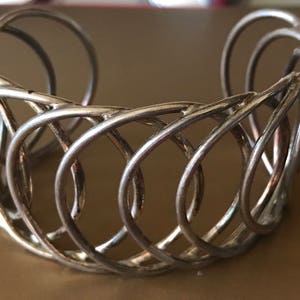 May include: A silver metal bracelet with a woven design. The bracelet is made of multiple intertwined loops of metal.