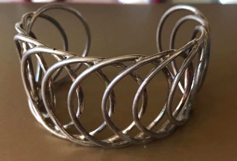 May include: A silver metal bracelet with a woven design. The bracelet is made of multiple intertwined loops of metal.