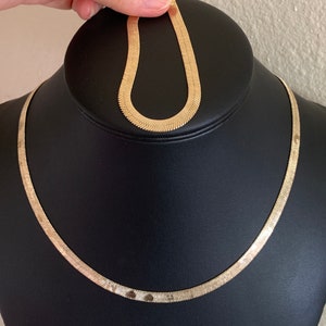 May include: A gold snake chain necklace on a black display stand. The chain is made of small, flat links that create a smooth, flowing look.