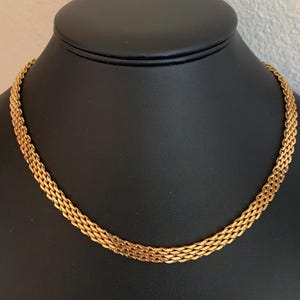 May include: A gold-colored necklace with a woven chain design. The necklace is displayed on a black mannequin. The chain is made of multiple strands woven together, creating a textured appearance. The necklace is a classic piece of jewelry.