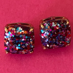 May include: A pair of square stud earrings featuring a gold-toned metal setting and a multi-colored glitter inlay. The surface of each earring is filled with reflective, sparkling flecks in shades of blue, red, pink, and silver, creating a dense, textured appearance.