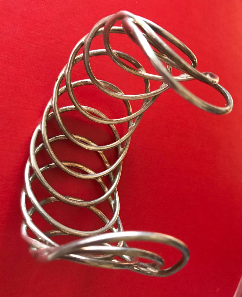May include: A silver metal bracelet with a unique, multi-layered design. The bracelet is made of several intertwined rings, creating a textured and intricate look.