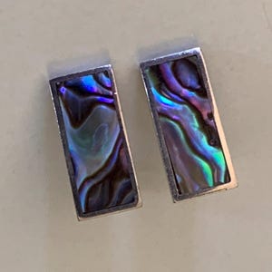 May include: A pair of rectangular earrings featuring iridescent abalone shell inlays set within polished silver-toned metal frames. The shell displays swirling patterns of deep purple, vibrant blue, and green hues across the flat, smooth surfaces.