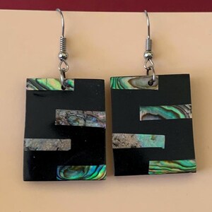 May include: A pair of rectangular earrings with a black base and inlaid iridescent abalone shell accents. The earrings have silver-colored fish hook ear wires. The abalone shell pieces display shades of green, blue, and silver.