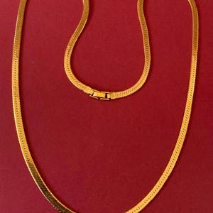 May include: A gold-colored herringbone chain necklace. The necklace is displayed against a red background. The chain has a flat, woven design and a clasp closure.