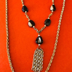 May include: A gold chain necklace with a black and white stone pendant and a tassel. The necklace has a rope chain style and is on a red background.