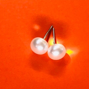 May include: A pair of pearl stud earrings with silver posts. The earrings are on a bright orange surface.