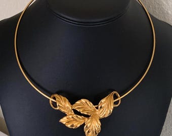May include: A gold-toned necklace with a leaf design. The necklace features a rigid, circular collar with three stylized leaves clustered at the front. The leaves have detailed veining and a polished finish, creating a classic and elegant look.