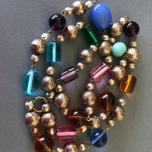 May include: A colorful beaded necklace with a mix of round and rectangular glass beads in shades of blue, green, purple, pink, and amber. The necklace is strung on a silver chain with decorative metal beads.