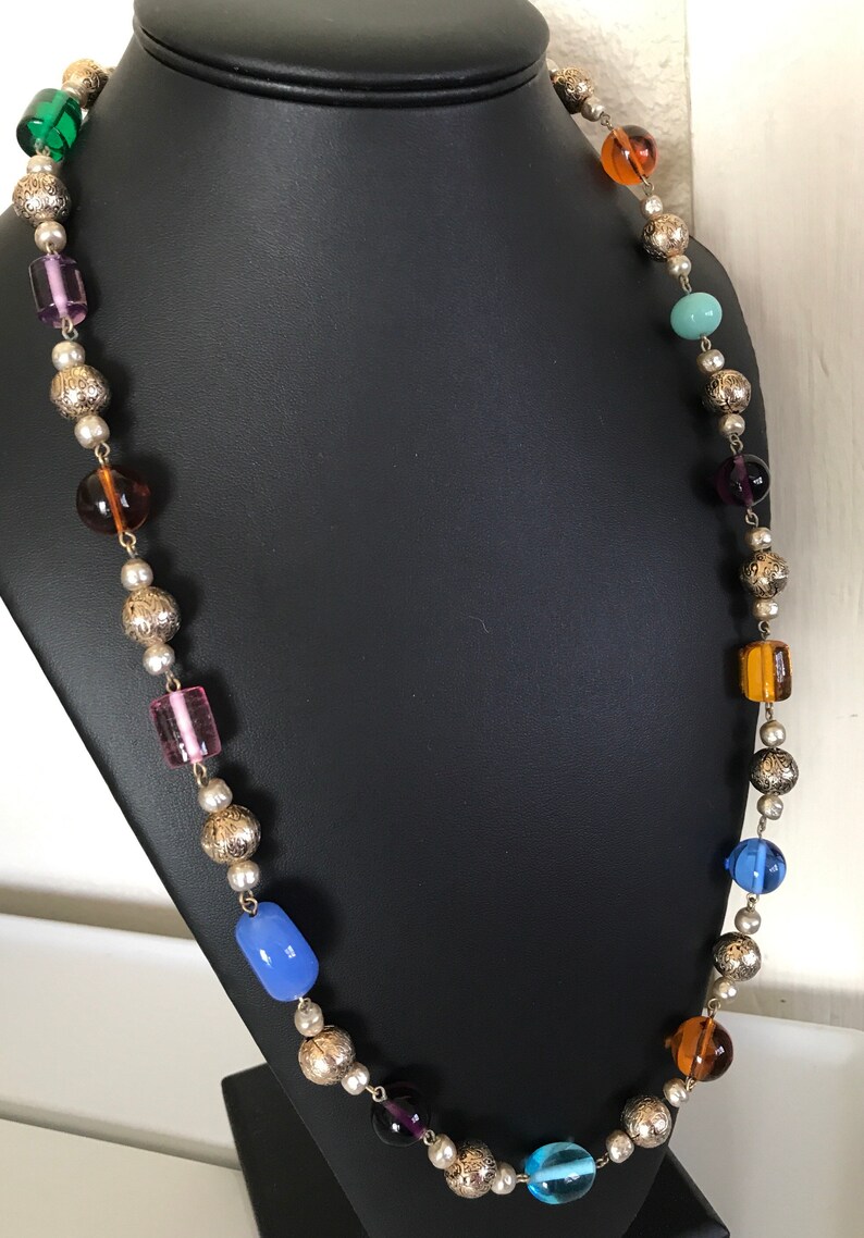 May include: A long necklace with silver beads and colorful glass beads. The beads are in various shapes and sizes, including round, square, and rectangular. The necklace is displayed on a black mannequin.