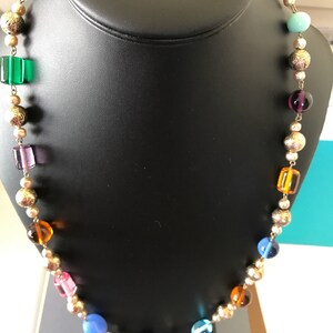 May include: A colorful beaded necklace with gold accents. The necklace features a variety of round and square beads in shades of blue, green, purple, orange, and pink.