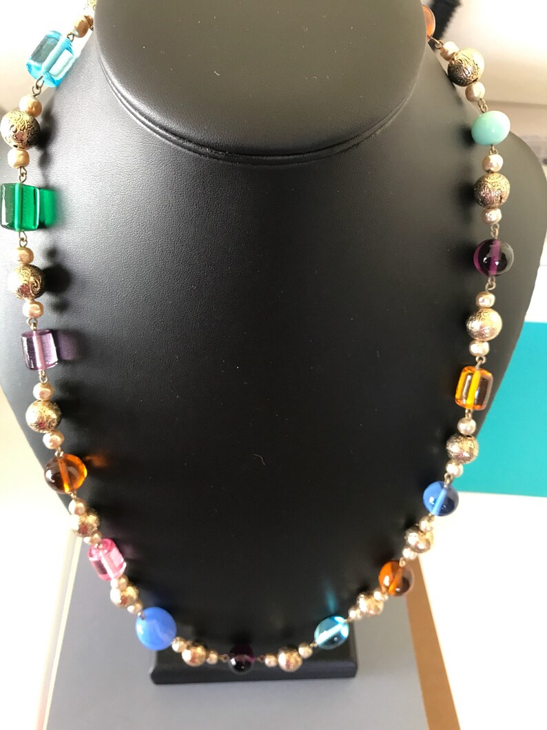 May include: A colorful beaded necklace with gold accents. The necklace features a variety of round and square beads in shades of blue, green, purple, orange, and pink.
