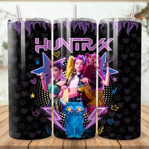 Puede incluir: Un vaso negro con tapa y pajita moradas. El vaso presenta un gr&aacute;fico de tres mujeres con micr&oacute;fonos, la palabra "HUNTRAX" y un gato azul. El fondo tiene estrellas, corazones y puntos.