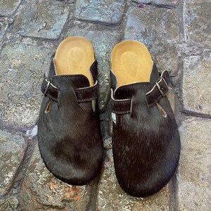 May include: A pair of dark brown clogs with a fur-like texture. The clogs have a light brown insole and a buckle strap. The shoes are displayed on a cobblestone surface.