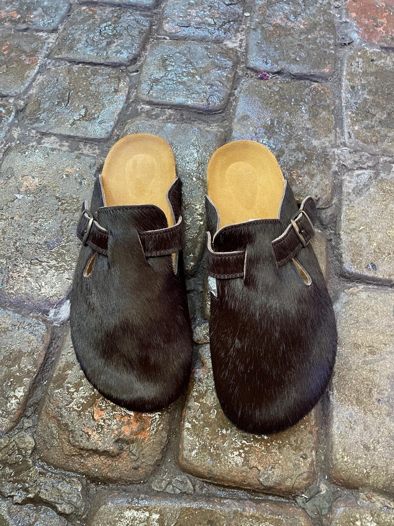 May include: A pair of dark brown clogs with a fur-like texture. The clogs have a light brown insole and a buckle strap. The shoes are displayed on a cobblestone surface.