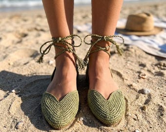 May include: A pair of olive green woven flats featuring a pointed toe with a V-shaped cutout and braided ankle ties that wrap around the lower leg. The shoes have a textured, cord-like construction and are designed to be tied securely at the ankle.