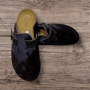 May include: A pair of black fur clogs with adjustable straps and buckle closures. The clogs have a contoured footbed and are set against a wooden background. The fur appears to be a dark, natural color.