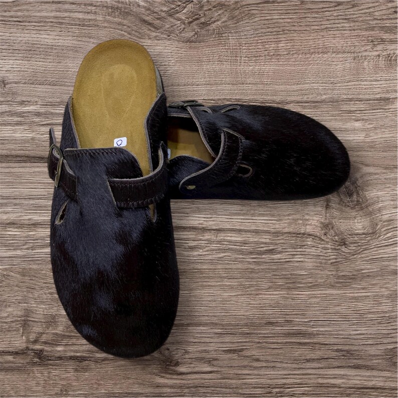 May include: A pair of black fur clogs with adjustable straps and buckle closures. The clogs have a contoured footbed and are set against a wooden background. The fur appears to be a dark, natural color.