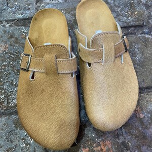 May include: A pair of tan-colored clogs with a fur-like texture. Each clog features a buckle closure and a contoured footbed. The clogs are designed for comfort and casual wear.