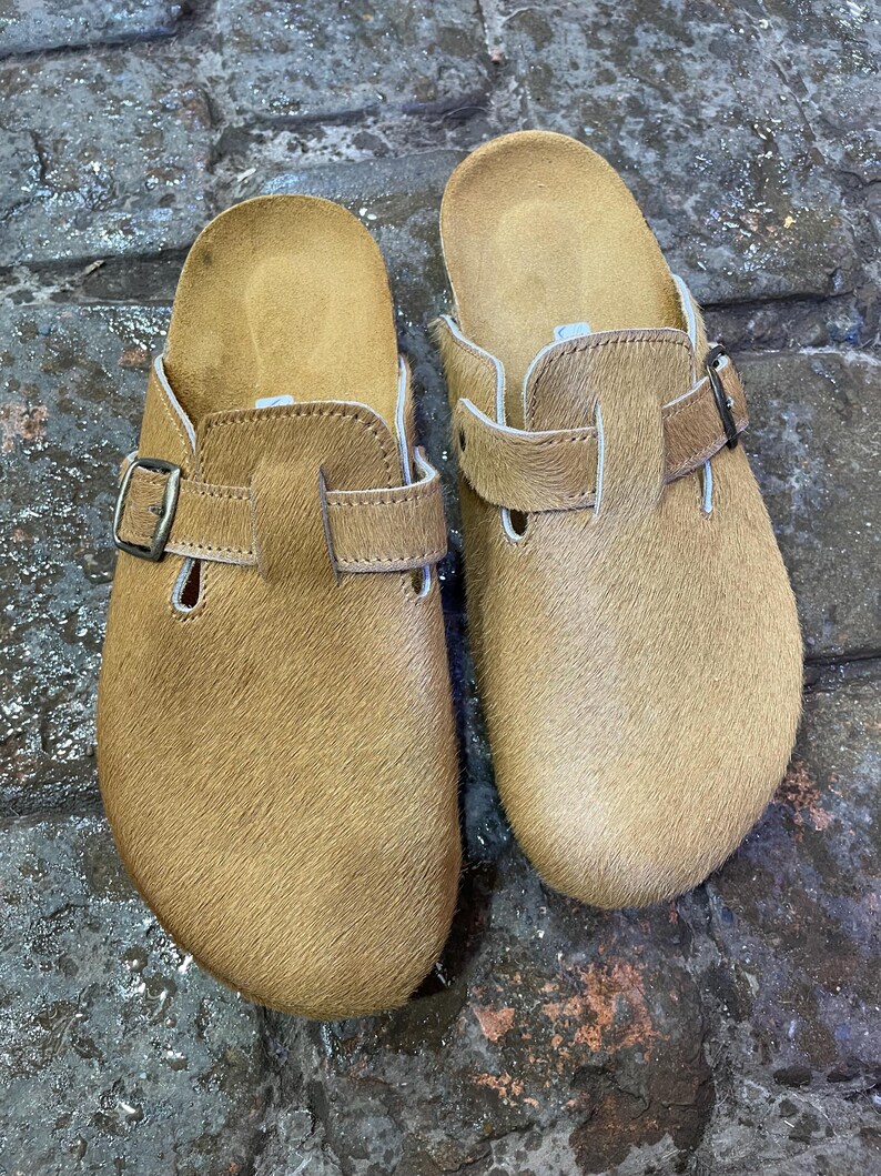 May include: A pair of tan-colored clogs with a fur-like texture. Each clog features a buckle closure and a contoured footbed. The clogs are designed for comfort and casual wear.