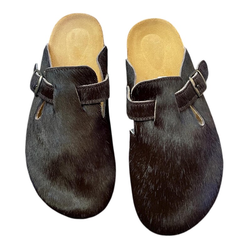 May include: A pair of black fur clogs with a cork footbed and adjustable buckle straps. The clogs have a dark brown fur exterior and a light brown interior. The buckles are silver in color.