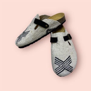 May include: A pair of off-white clogs with black detailing. The clogs have a woven texture, black embroidered patterns, and black straps with buckles. The soles are cork and the background is a light pink.