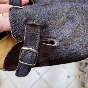 May include: Close-up of a pair of dark brown leather gloves with a textured, fur-like appearance. The gloves feature a buckle closure and a strap detail. The leather has a rich, dark color.