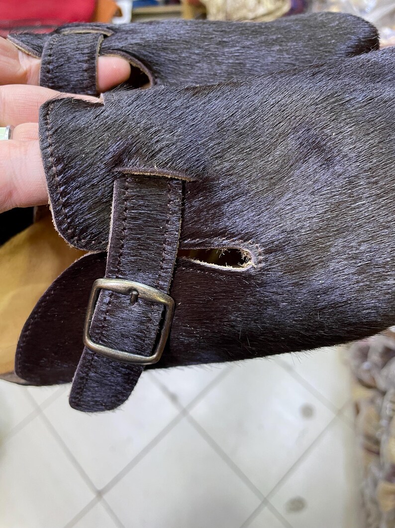 May include: Close-up of a pair of dark brown leather gloves with a textured, fur-like appearance. The gloves feature a buckle closure and a strap detail. The leather has a rich, dark color.