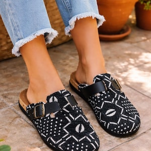 May include: Black and white patterned clogs with a cork footbed and black leather straps. The clogs have a buckle closure and a geometric design. The shoes are worn with light blue denim jeans with frayed edges.