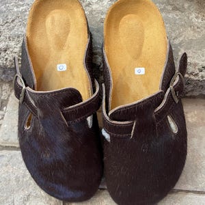 May include: A pair of brown leather clogs with a fur-like texture. The clogs have a buckle closure and a tan-colored insole. The shoes are designed with an open-back style, suitable for casual wear.