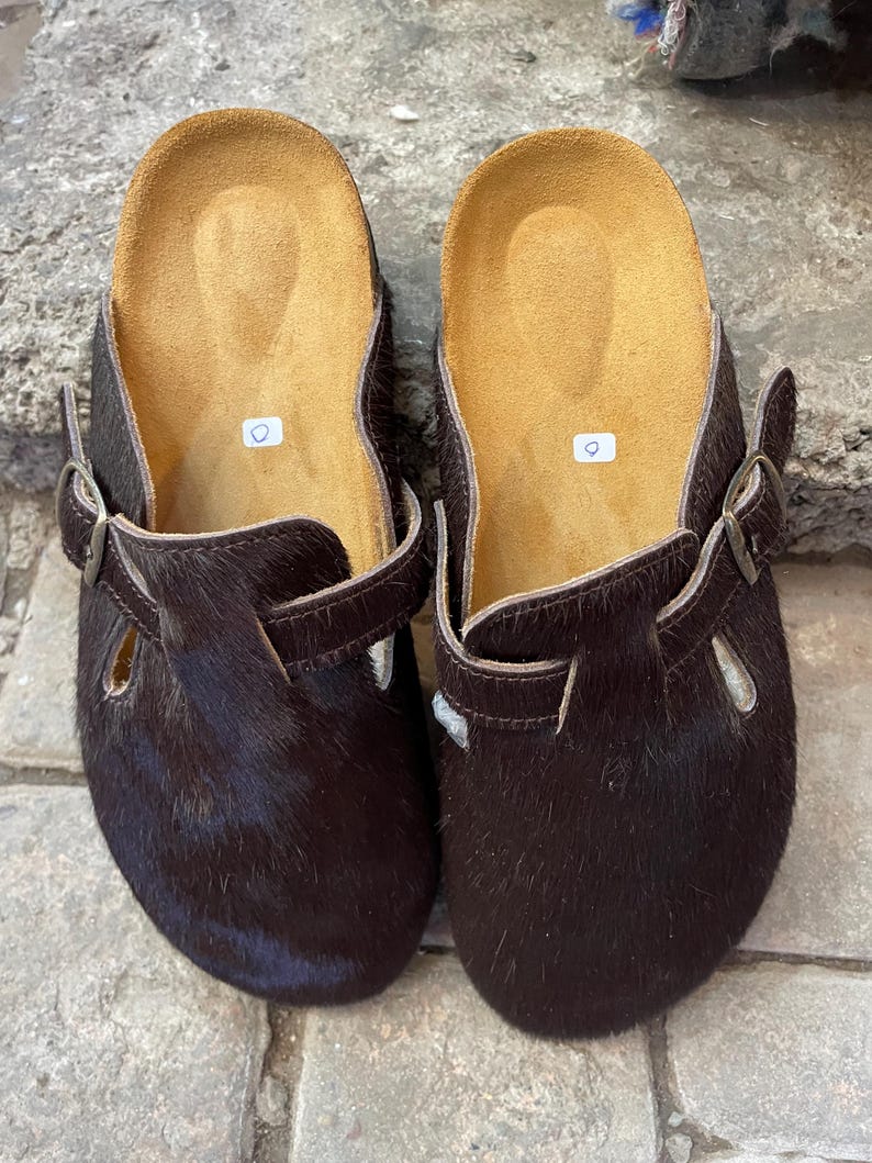 May include: A pair of brown leather clogs with a fur-like texture. The clogs have a buckle closure and a tan-colored insole. The shoes are designed with an open-back style, suitable for casual wear.