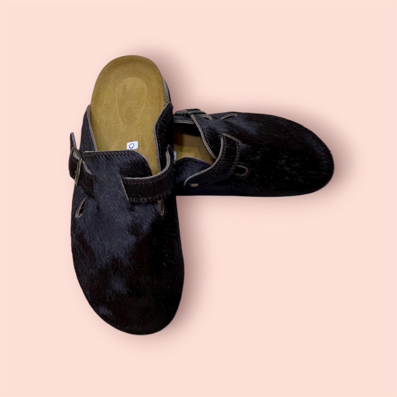 May include: A pair of black clogs with a fur-like texture. The clogs have a brown footbed and adjustable straps with buckles. The shoes are set against a light pink background.