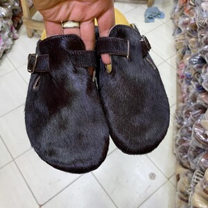 May include: A pair of black fur clogs with leather straps and buckle closures. The clogs have a rounded toe and a dark brown leather sole. The shoes are held up by a hand.