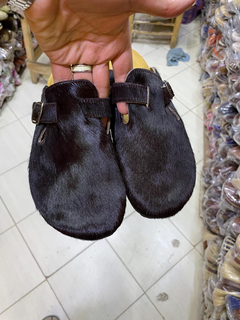 May include: A pair of black fur clogs with leather straps and buckle closures. The clogs have a rounded toe and a dark brown leather sole. The shoes are held up by a hand.