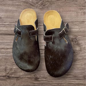 May include: A pair of black clogs with a fur-like texture. The clogs have a brown leather strap with a silver buckle and a light brown insole. The shoes are displayed on a wooden surface.