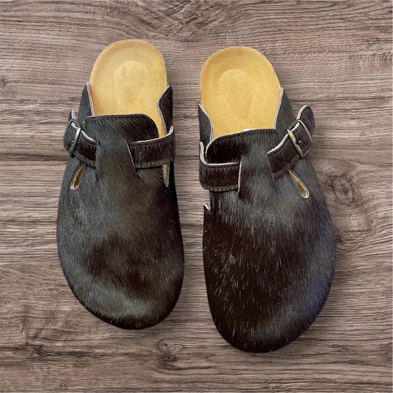 May include: A pair of black clogs with a fur-like texture. The clogs have a brown leather strap with a silver buckle and a light brown insole. The shoes are displayed on a wooden surface.