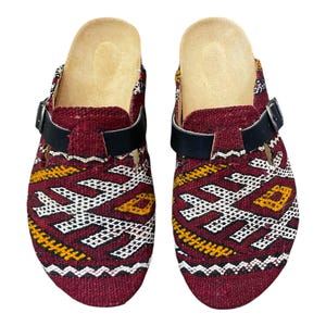 Könnte beinhalten: Paar weinrote Clogs mit einem gewebten Muster in Weiß, Gelb und Schwarz. Die Clogs haben einen schwarzen Lederriemen mit Schnalle und eine beige Innensohle. Diese Schlupfschuhe sind ein einzigartiger Stil.