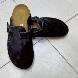 May include: Black fur clogs with brown leather straps and buckles. The clogs have a contoured footbed and a slip-on design. The shoes are made of a dark, textured material and are designed for comfort and style.