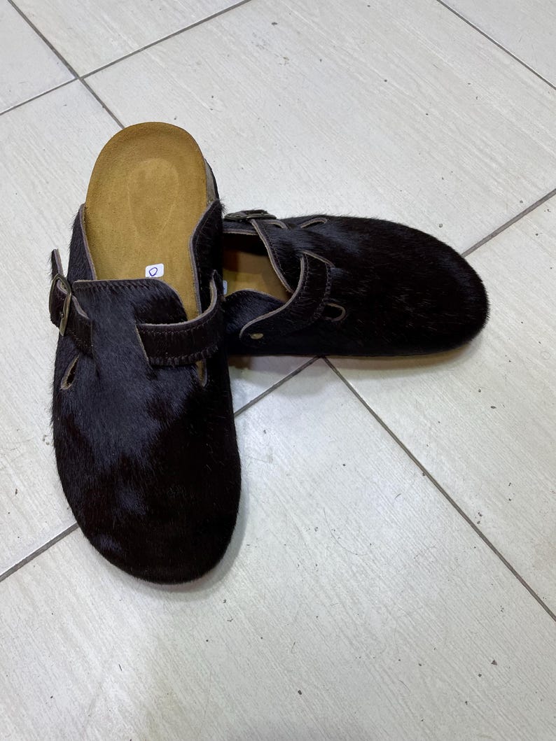 May include: Black fur clogs with brown leather straps and buckles. The clogs have a contoured footbed and a slip-on design. The shoes are made of a dark, textured material and are designed for comfort and style.