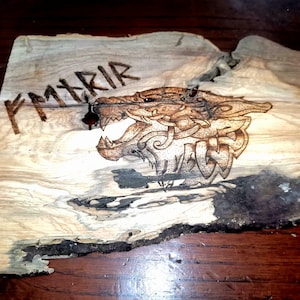 Puede incluir: Placa de madera con un diseño pirograbado de la cabeza de un lobo gruñendo. La obra de arte está en tonos marrones y negros, con la palabra "FMXRIR" encima del lobo. La madera tiene una forma natural e irregular.