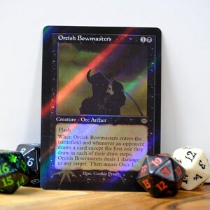 May include: A holographic Orcish Bowmasters trading card with a silhouetted archer image. The card features text describing its abilities and is surrounded by various dice, including a green and black D20, and a black and red D12.