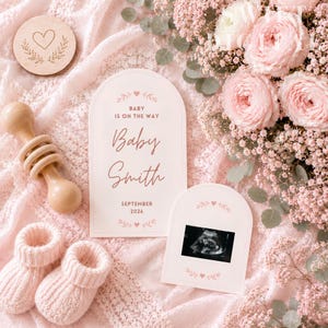 Peut inclure: Deux cartes d'annonce arquées avec du texte rose indiquant Baby Is On The Way, Baby Smith et September 2026, accompagnées d'un hochet en bois, de chaussons tricotés, d'un disque en bois gravé d'un cœur et d'un bouquet de roses roses.