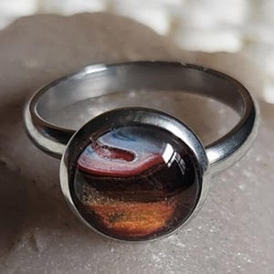 May include: A silver ring with a large, round, cabochon-style stone. The stone features a colorful abstract design with shades of brown, red, blue, and orange. The ring band is a simple, polished metal.