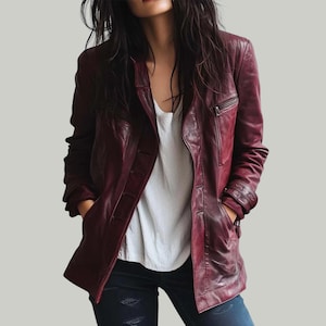 May include: A burgundy leather jacket with a button-up front, a zippered chest pocket, and two front pockets. The jacket is worn over a white top and dark blue jeans.