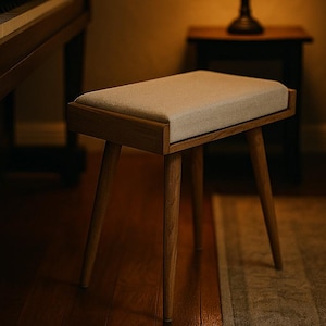 Puede incluir: Un taburete de piano de madera con un asiento rectangular acolchado en beige claro. Tiene un marco de madera y cuatro patas anguladas. El taburete est&aacute; en una habitaci&oacute;n con un piano, una l&aacute;mpara y una alfombra.