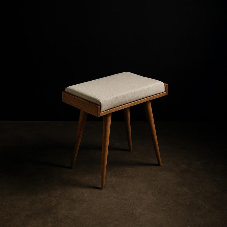 Puede incluir: Un peque&ntilde;o taburete de madera con un asiento rectangular y acolchado. El asiento es de color beige claro y el marco es de madera natural. El taburete tiene cuatro patas anguladas y el dise&ntilde;o general es minimalista y moderno.