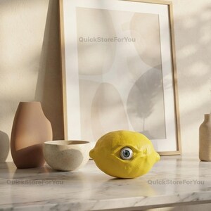 May include: A bright yellow lemon-shaped object featuring a single realistic human eye embedded in its center. The lemon has a textured surface and is positioned in front of a framed abstract print, a beige vase, and a speckled bowl.