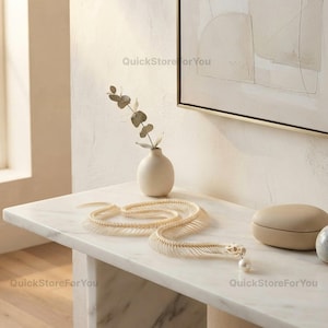 May include: A white snake skeleton sculpture with a single white pearl hanging from its jaw, positioned next to a small cream-colored vase containing a sprig of eucalyptus and a smooth, oval-shaped beige stone object on a white marble surface.