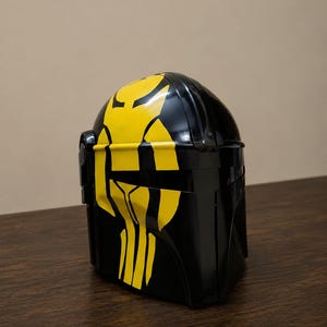 May include: A glossy black helmet featuring a prominent yellow graphic design on the front. The helmet has a smooth, curved surface with a horizontal visor opening and a distinct, angular shape that covers the head and face area completely.